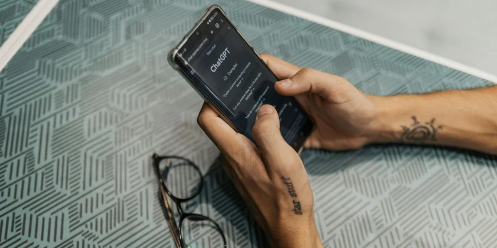 A person holding a smartphone with ChatGPT open, testing functionality on a different device, placed on a patterned surface.